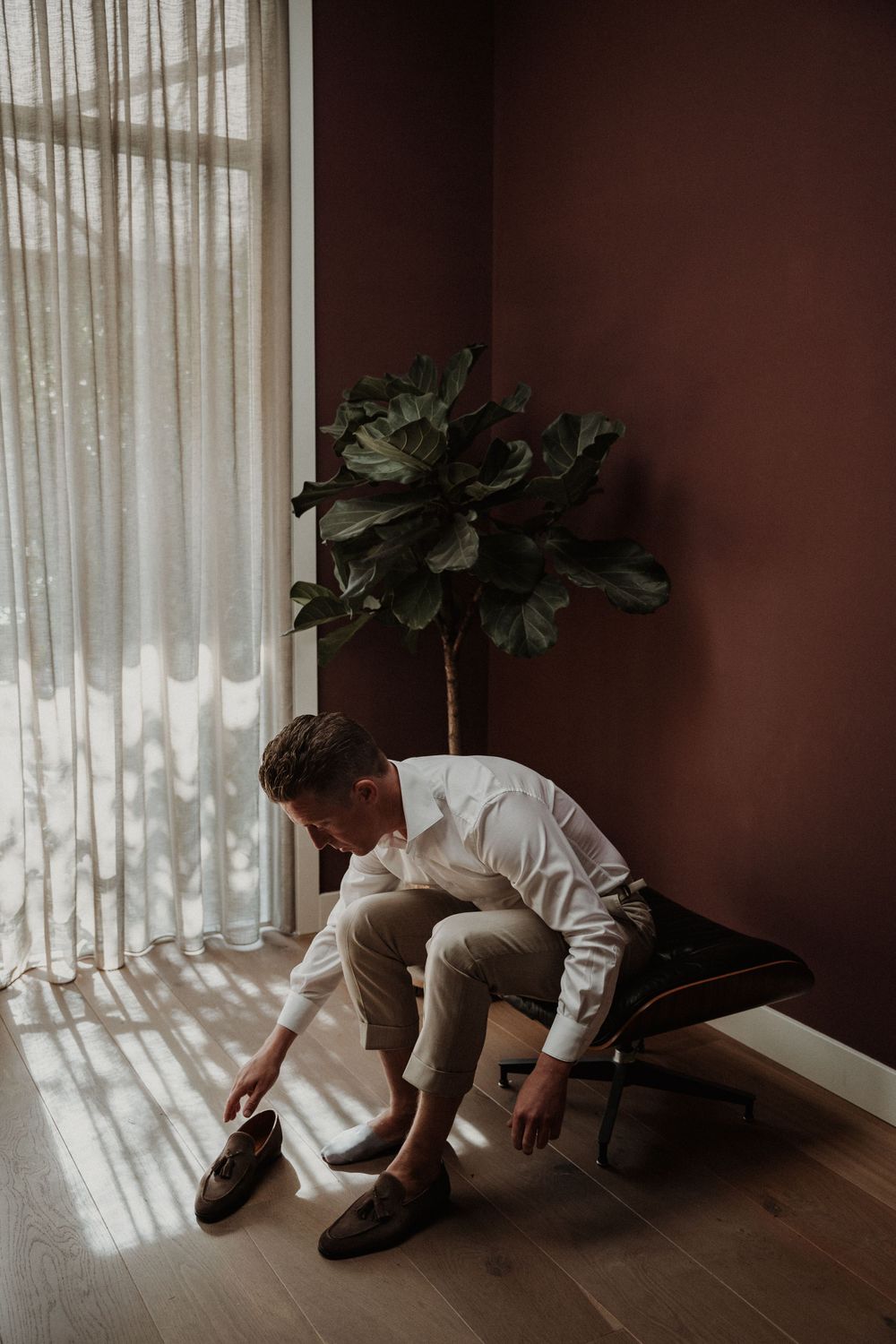 Someone in a white sweater and khaki pants puts on shoes near a window with soft natural light streaming through sheer curtains.