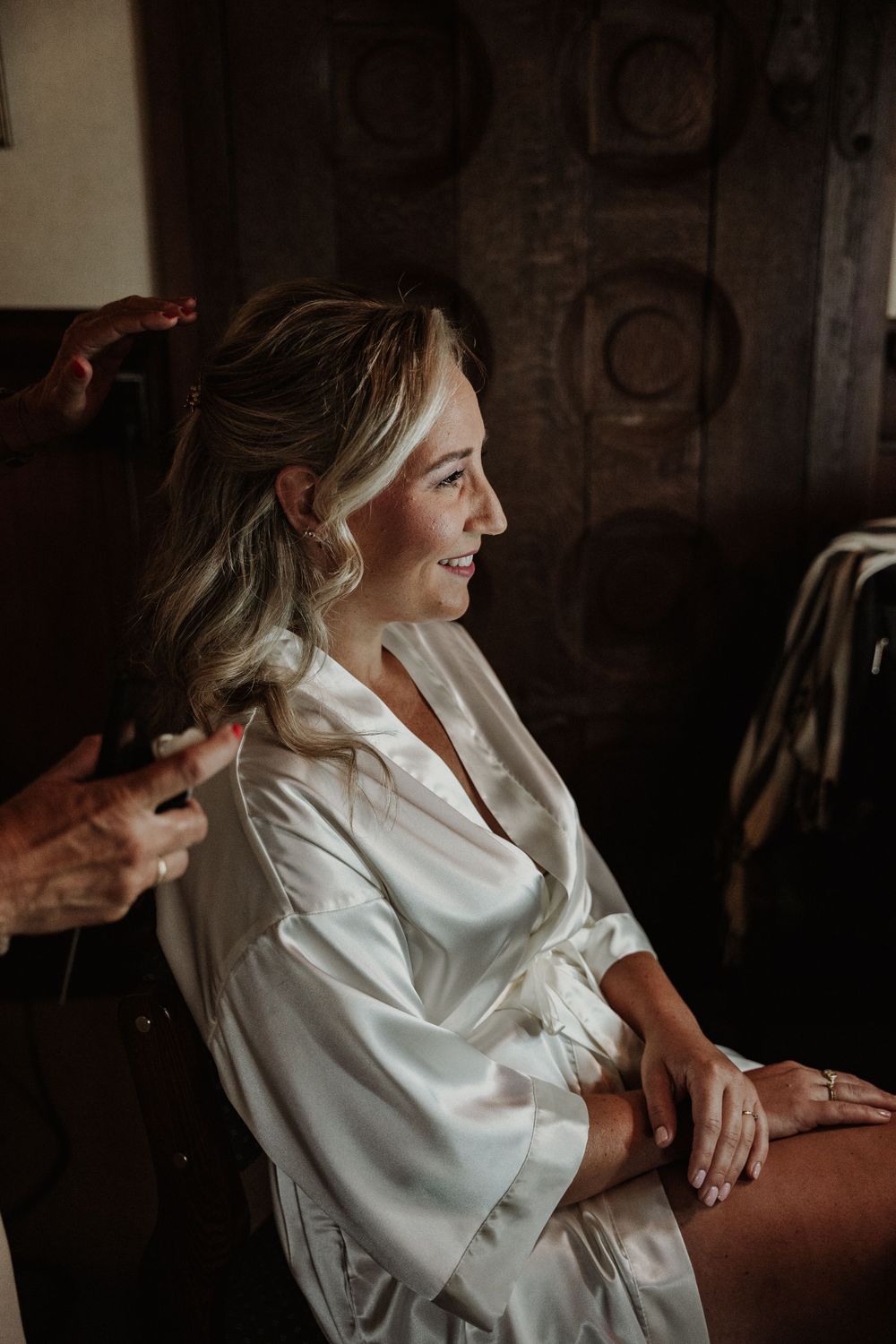 A person in a white silk robe sits while getting their hair styled in a dimly lit room.
