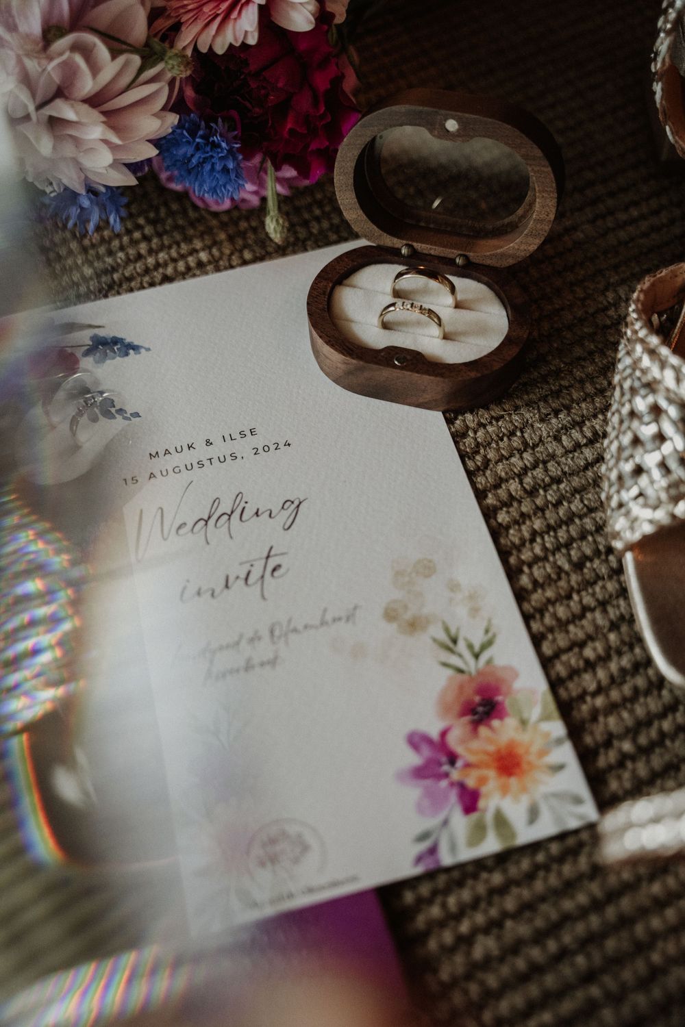 A wedding invitation with floral design lies next to a wooden ring box on a textured carpet.