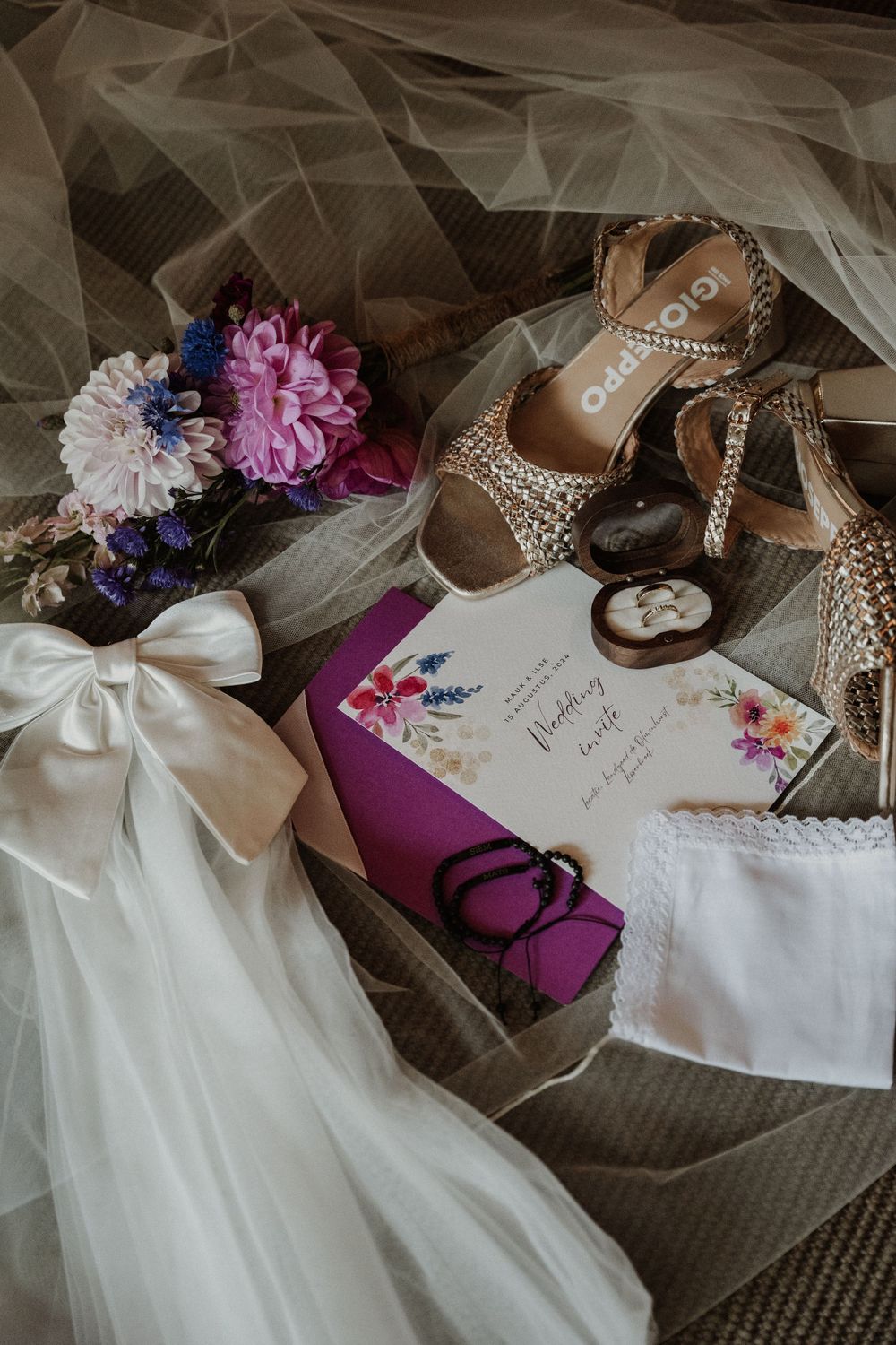 Wedding details including sparkly heels, floral bouquet, purple invitation, and white veil arranged on textured surface.