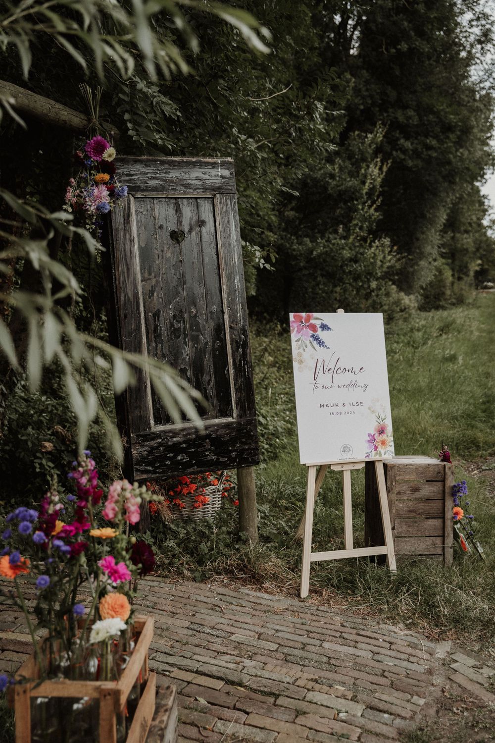 Rustic wedding welcome sign display with vintage door, wooden easel and colorful floral arrangements on brick path.