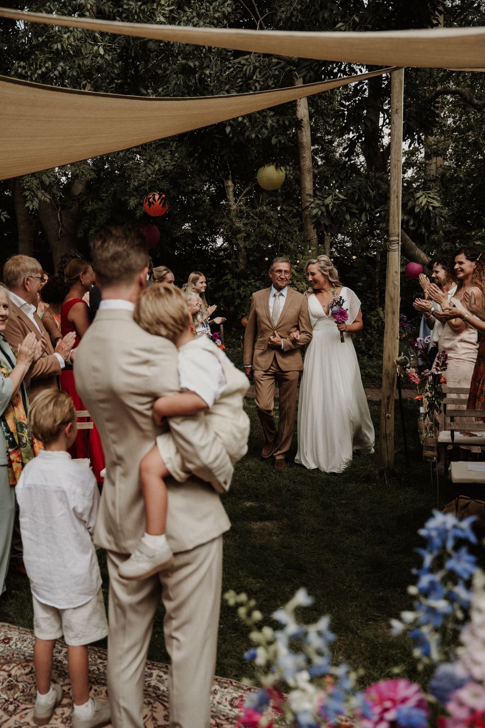 Outdoor garden wedding ceremony under tan canopies with guests gathered to celebrate the couple's special moment.