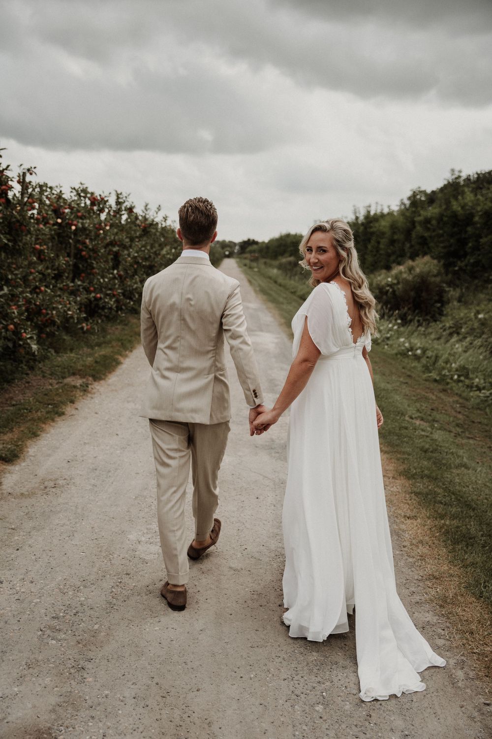 A couple in wedding attire walking down a dirt path surrounded by greenery on an overcast day.