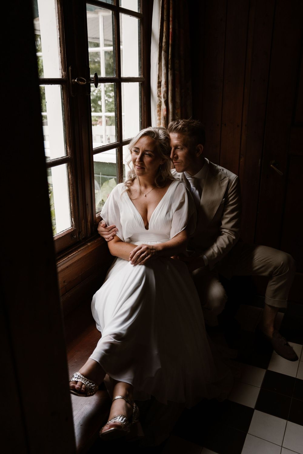 A romantic couple in wedding attire embraces by a window in moody lighting on a checkered floor.
