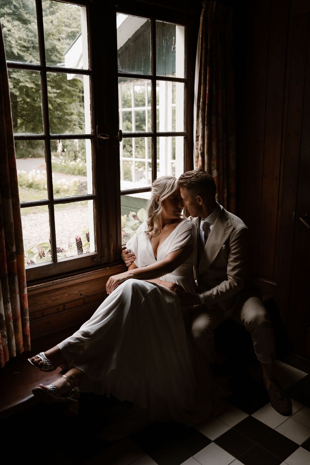 A romantic couple in wedding attire sits intimately by a rustic window with natural light streaming in.