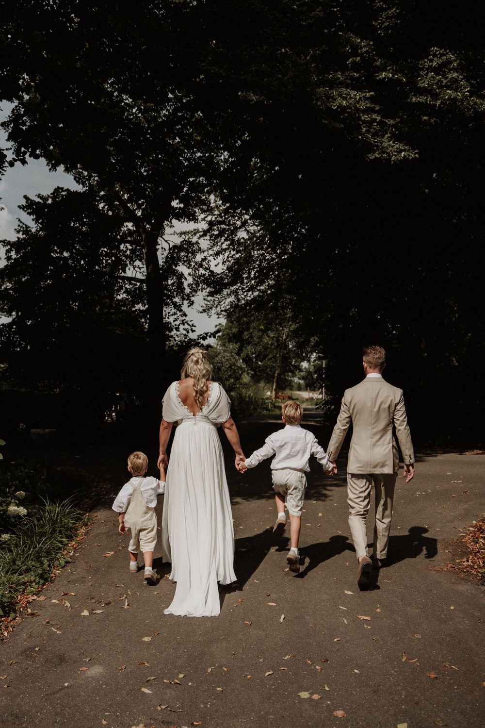 A family in wedding attire walks down a tree-lined path together on a sunny day.