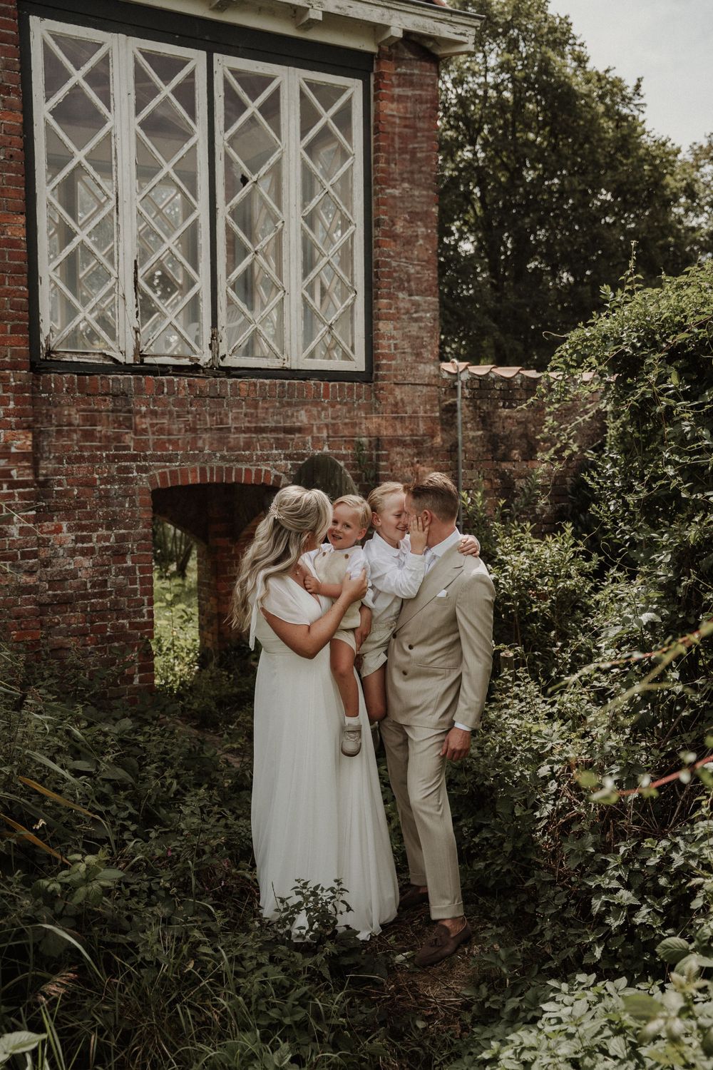A couple in wedding attire shares an intimate moment against a rustic brick building with white latticed windows.