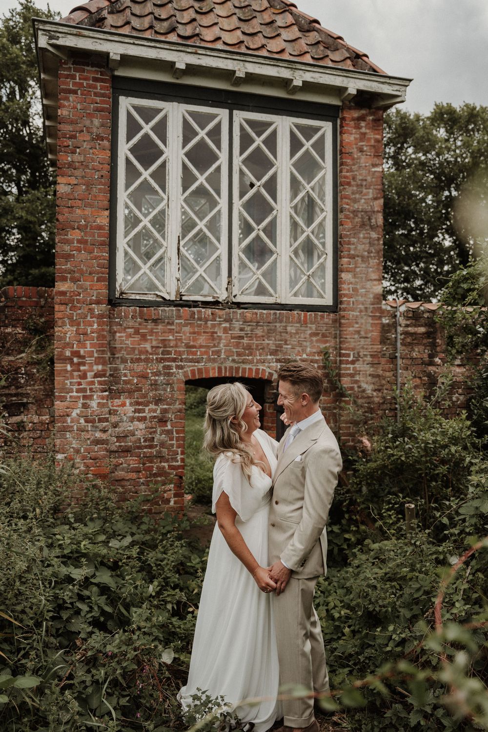 A romantic wedding couple embraces in front of a rustic brick building with diamond-paned windows.