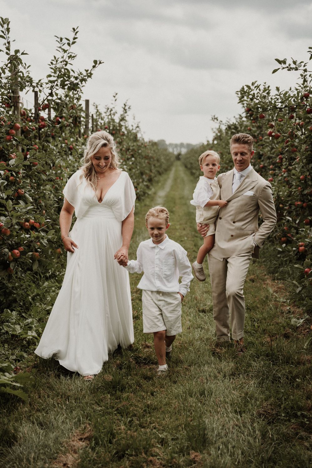 A family dressed in white and beige poses together in an apple orchard with rows of fruit trees on a cloudy day.