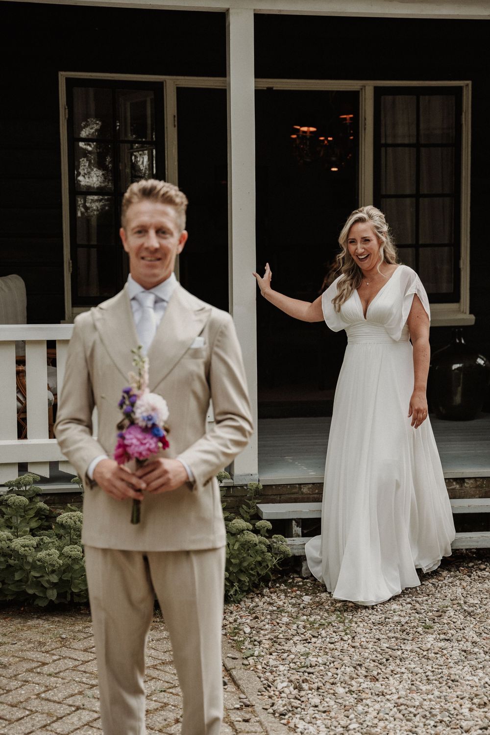A couple in wedding attire poses on a porch, with the bride in a flowing white dress and groom in a beige suit.
