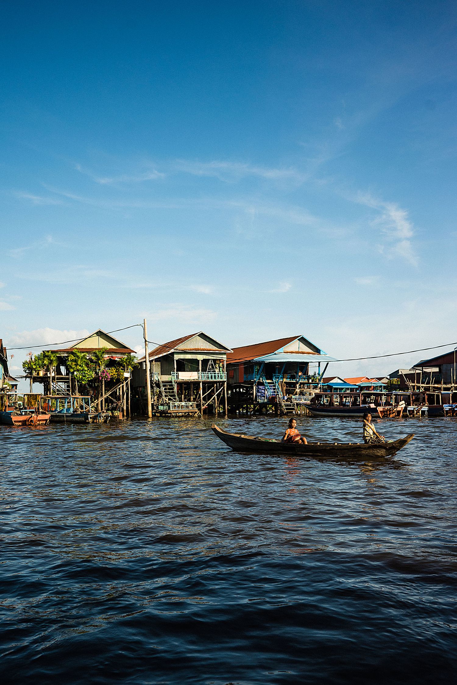 Wooden stilt houses along a river with small boats passing by in Southeast Asia.