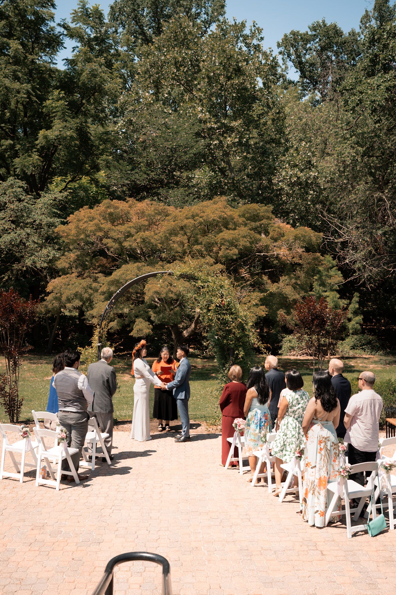A romantic couple embraces while standing on a wooden bridge with white railings surrounded by lush green trees.