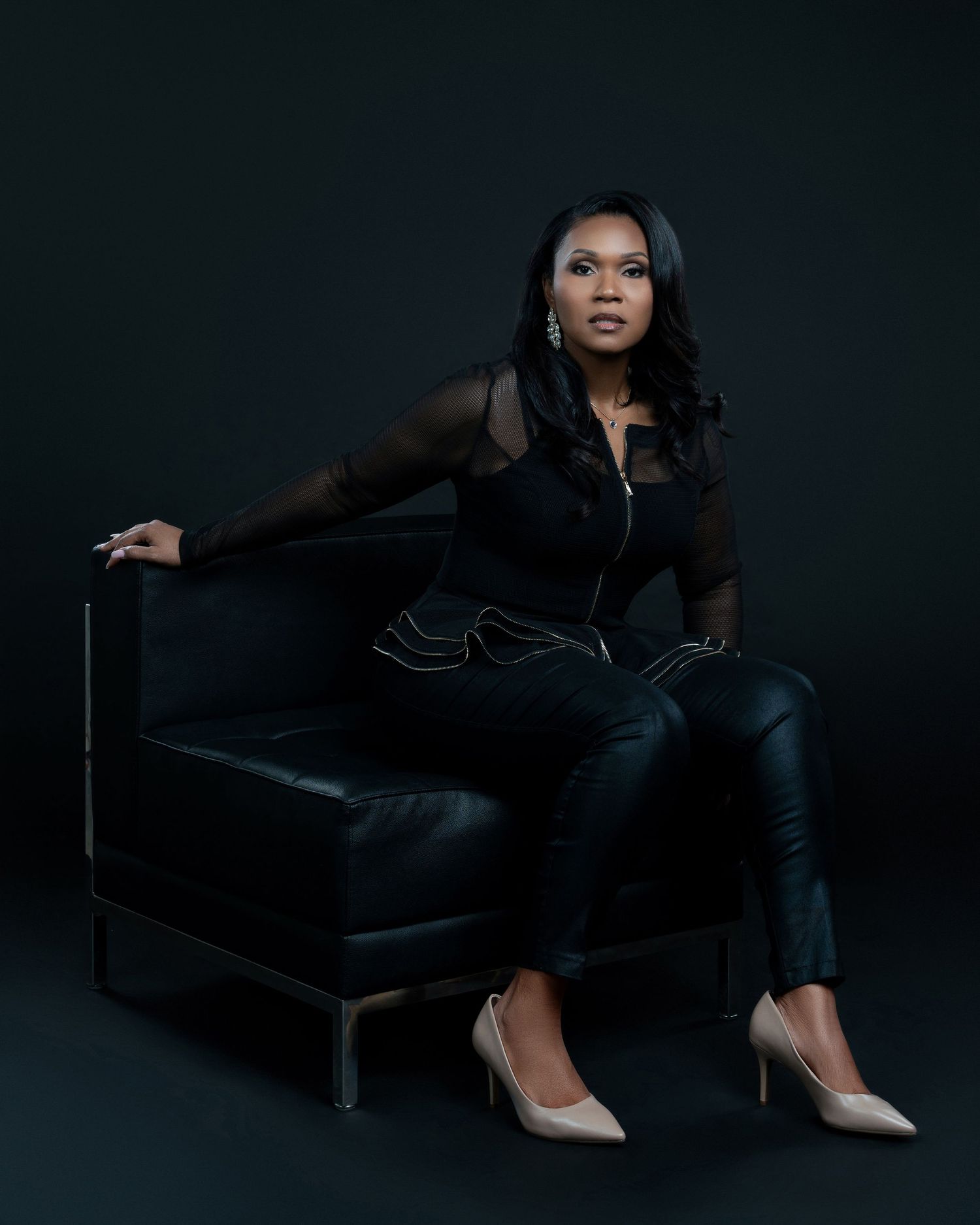 Stylish portrait of woman in black outfit seated on leather couch against dark backdrop.