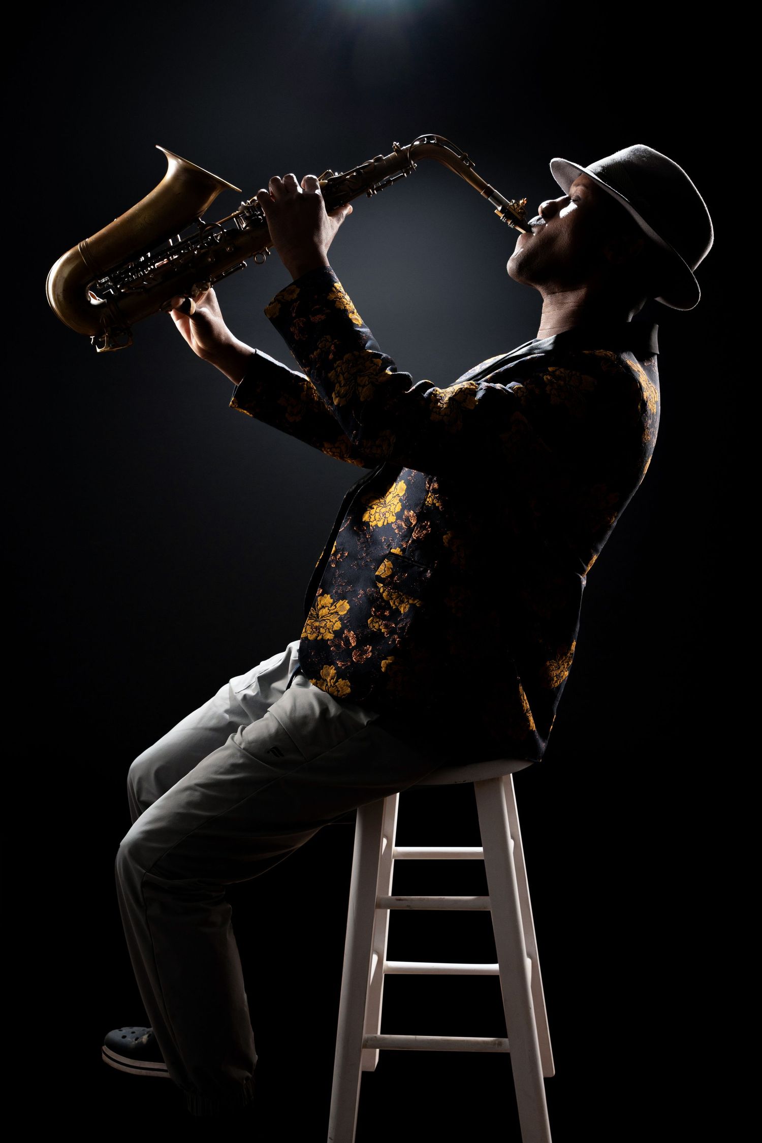 Dramatic side profile of musician playing saxophone while seated on stool in dark setting.