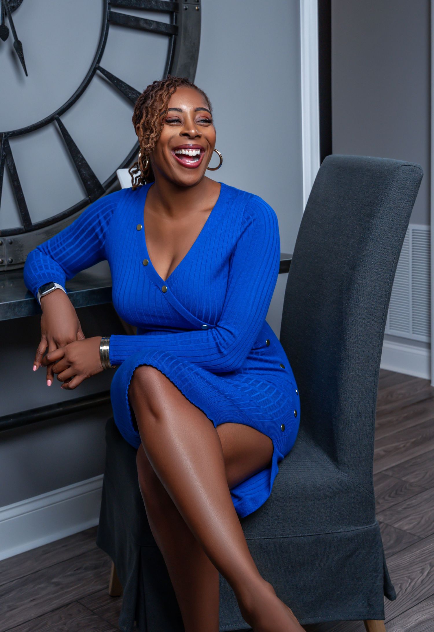 Professional portrait of someone in a vibrant blue wrap dress seated in a grey chair with a large clock on the wall.