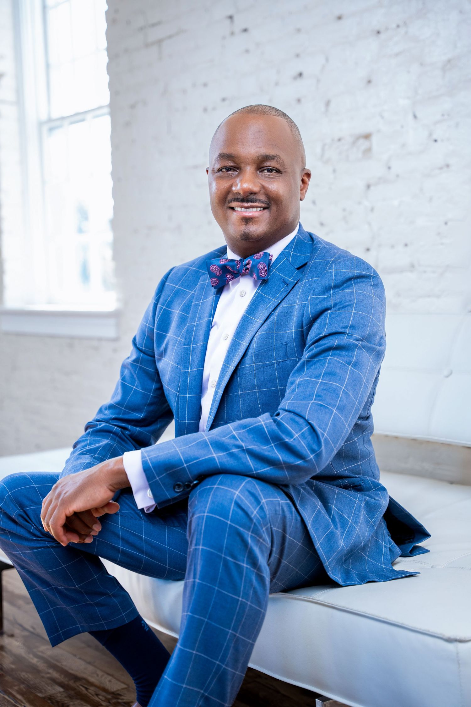 Professional in blue checkered suit and bow tie seated confidently on white bench.