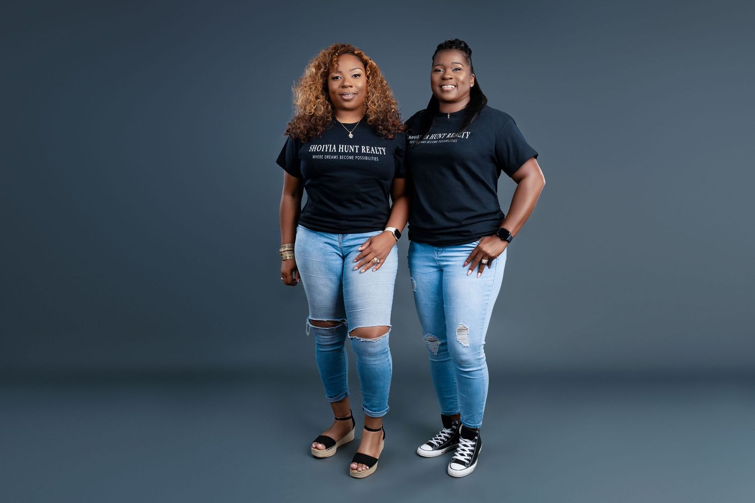 Two people in matching black t-shirts and distressed jeans pose against grey backdrop.