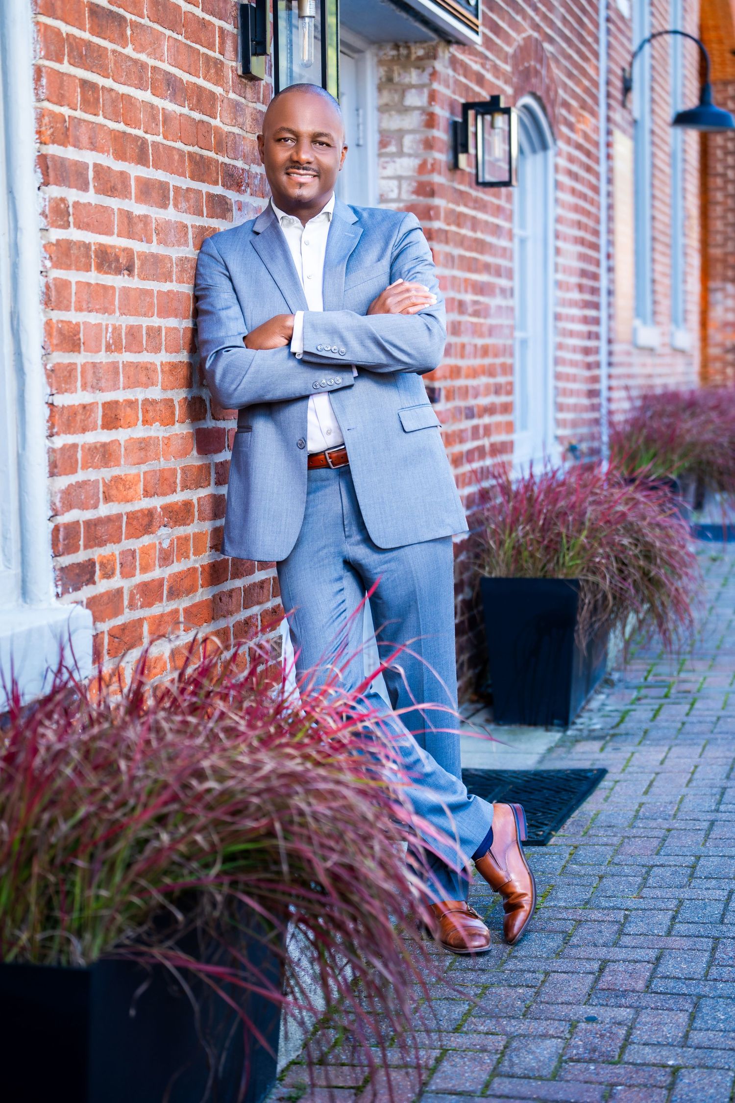 Business professional in light blue suit poses confidently against brick wall with decorative plants.