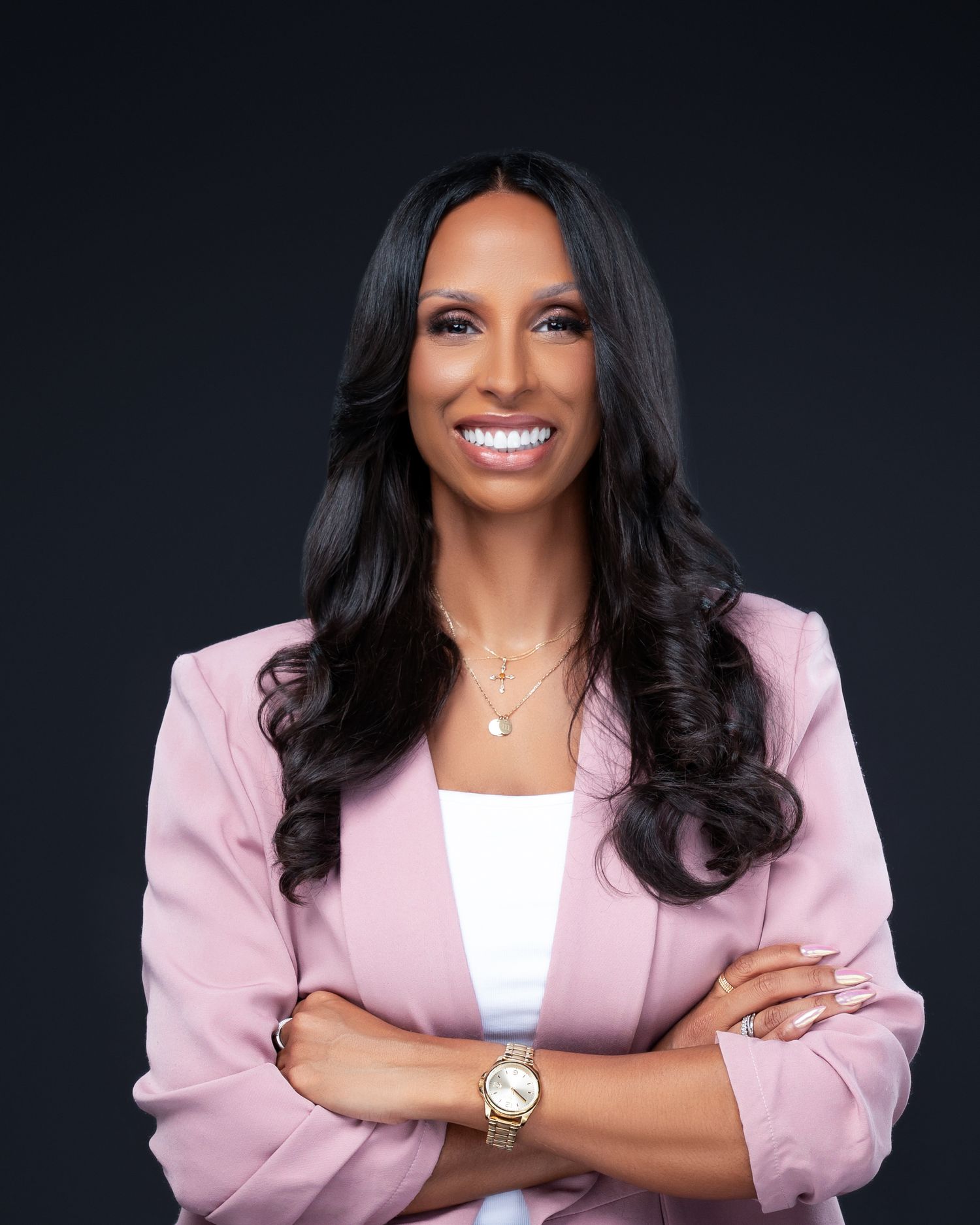 Professional woman in pink blazer and white top poses with crossed arms against dark backdrop.
