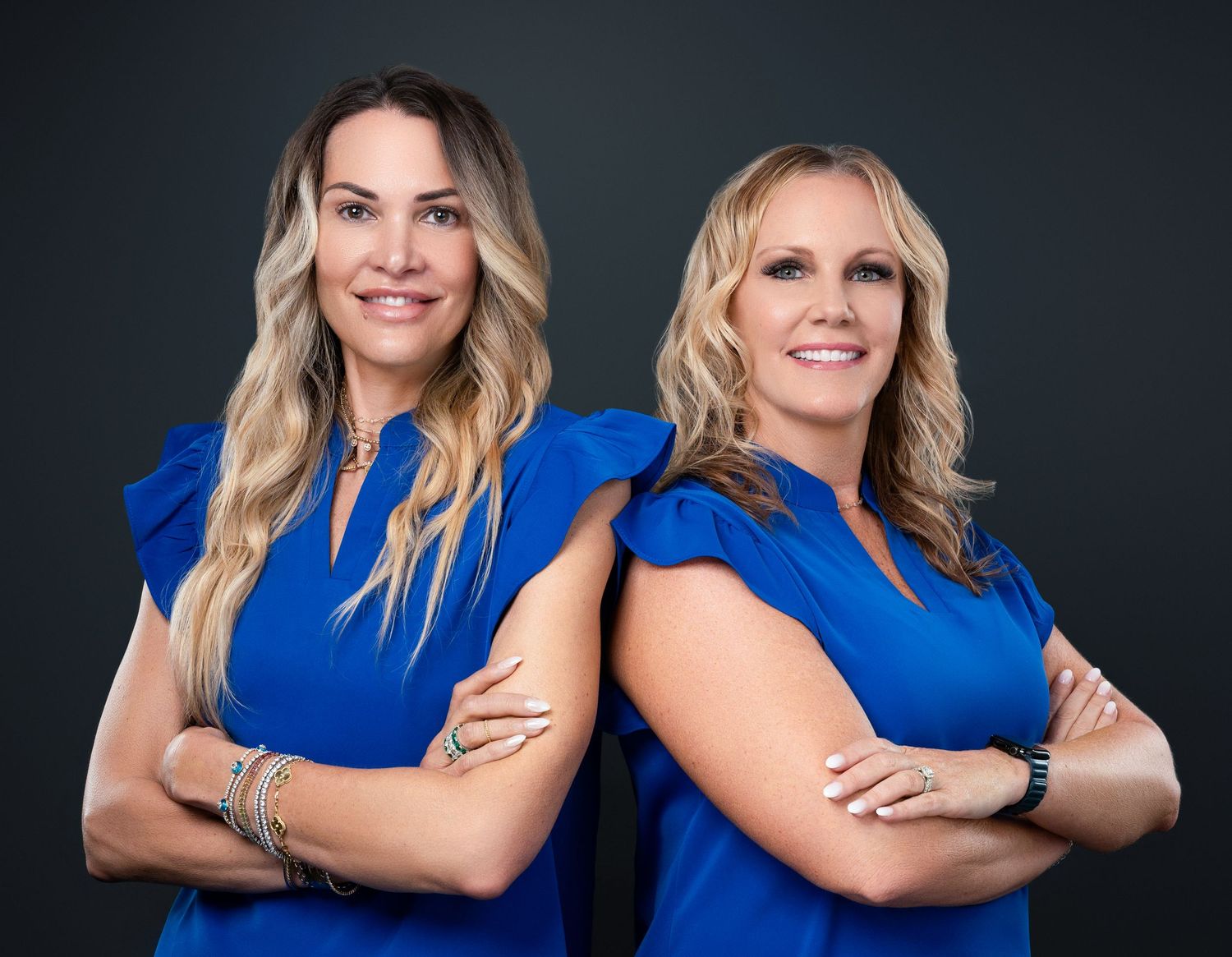 Two professionals in matching blue outfits pose confidently with crossed arms against dark background.