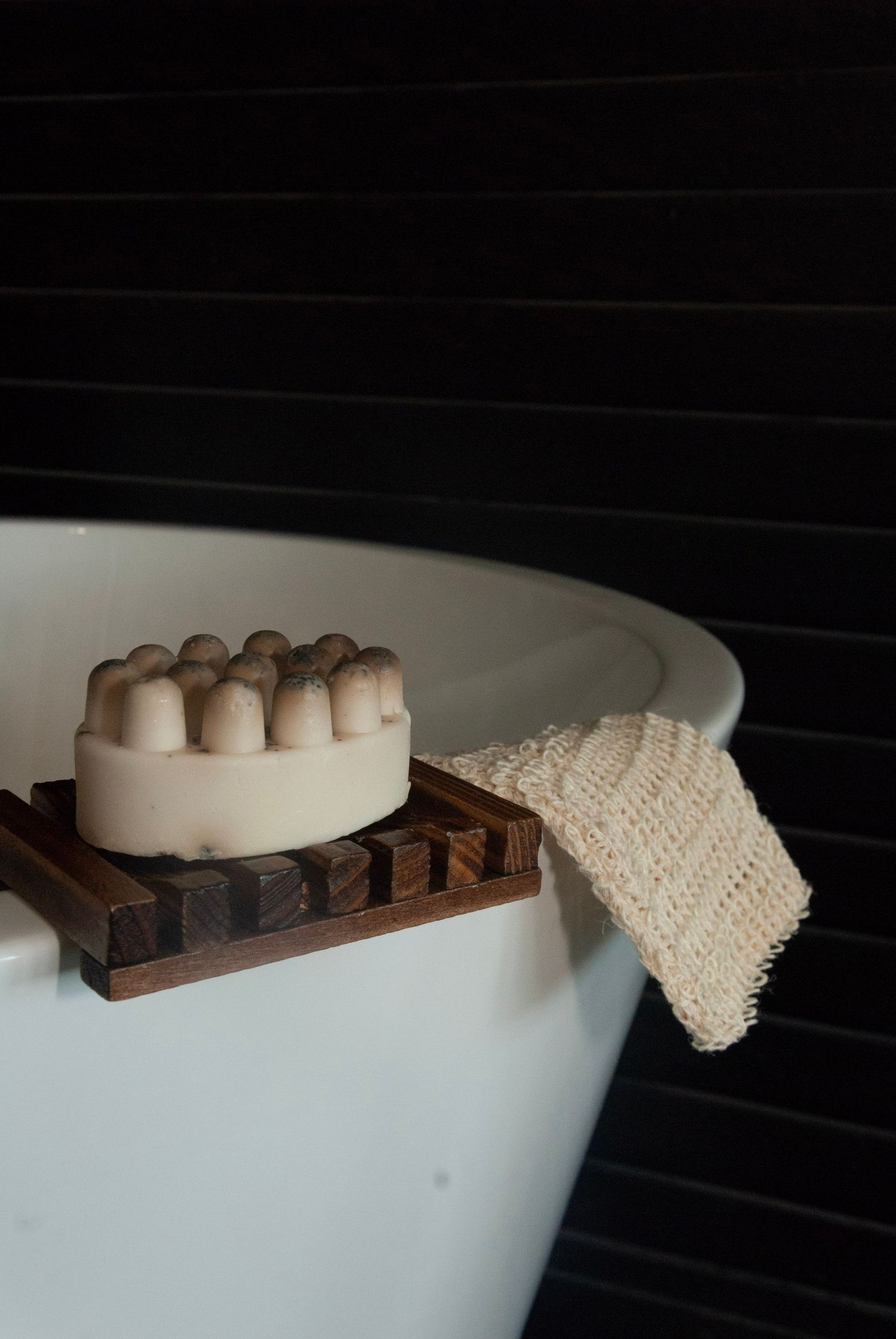 Bar of natural soap on wooden holder with knitted washcloth on edge of white bathtub.