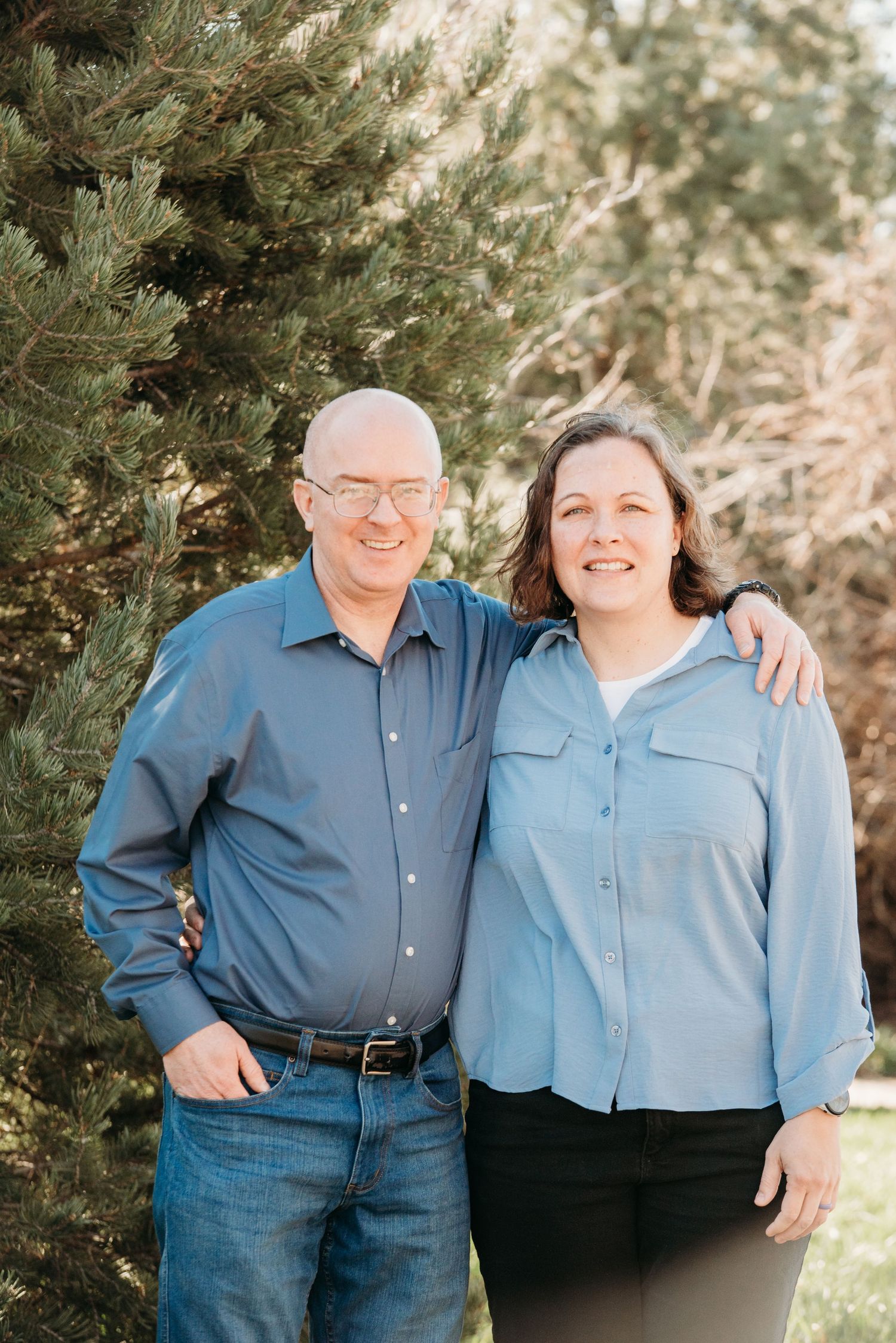 A couple stands together smiling in coordinated blue shirts against a backdrop of pine trees.