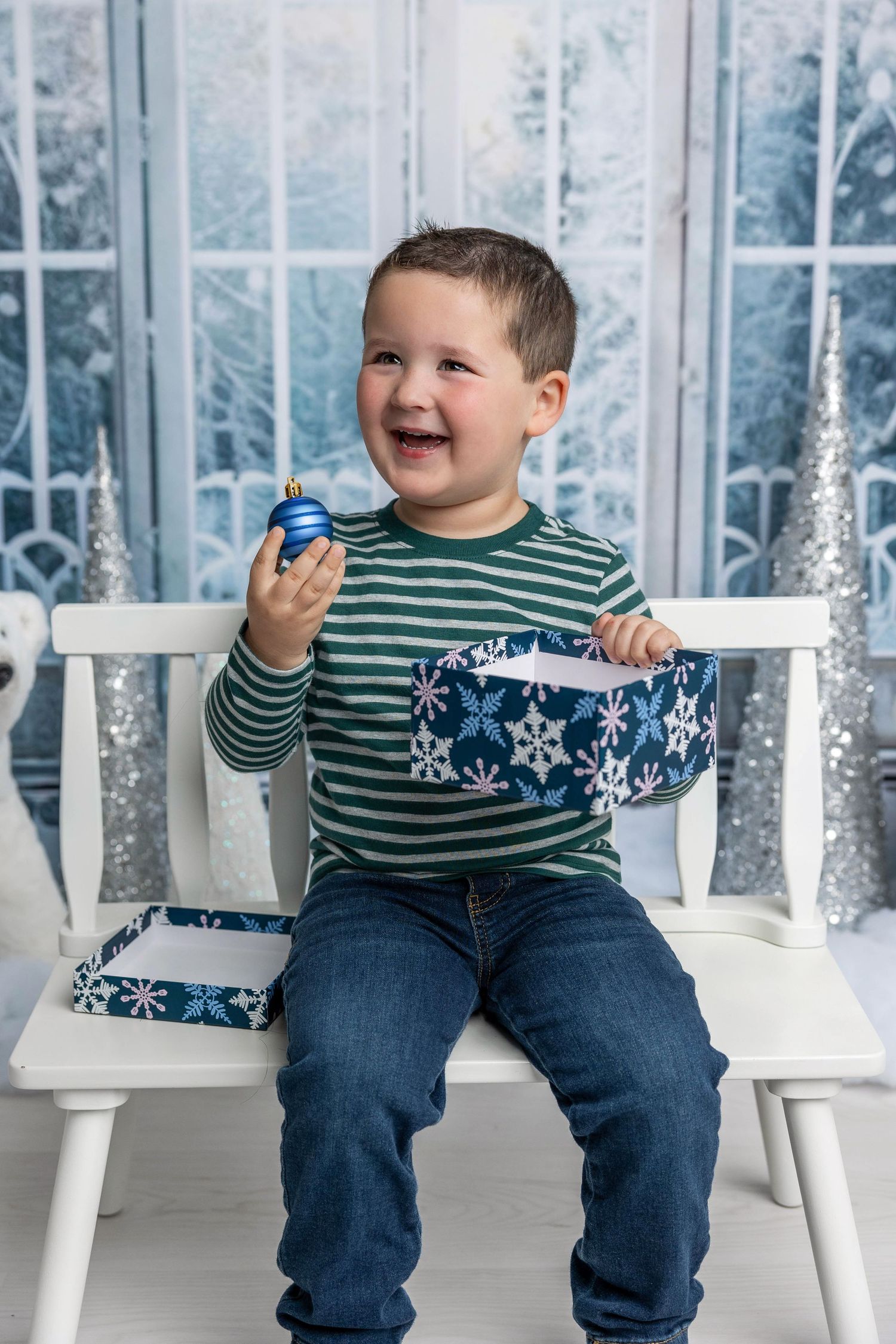 Child in green striped shirt sits on white bench with blue snowflake gift box during winter photo session.