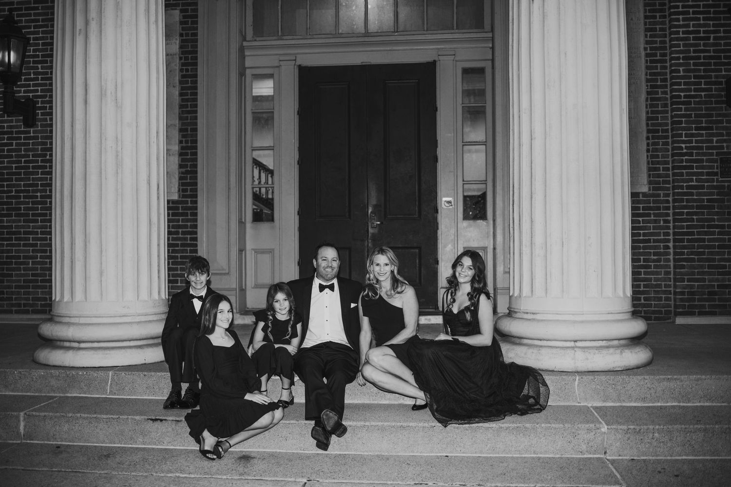 Black and white elegant formal photo of people sitting on the steps between white columns of a classic brick building.