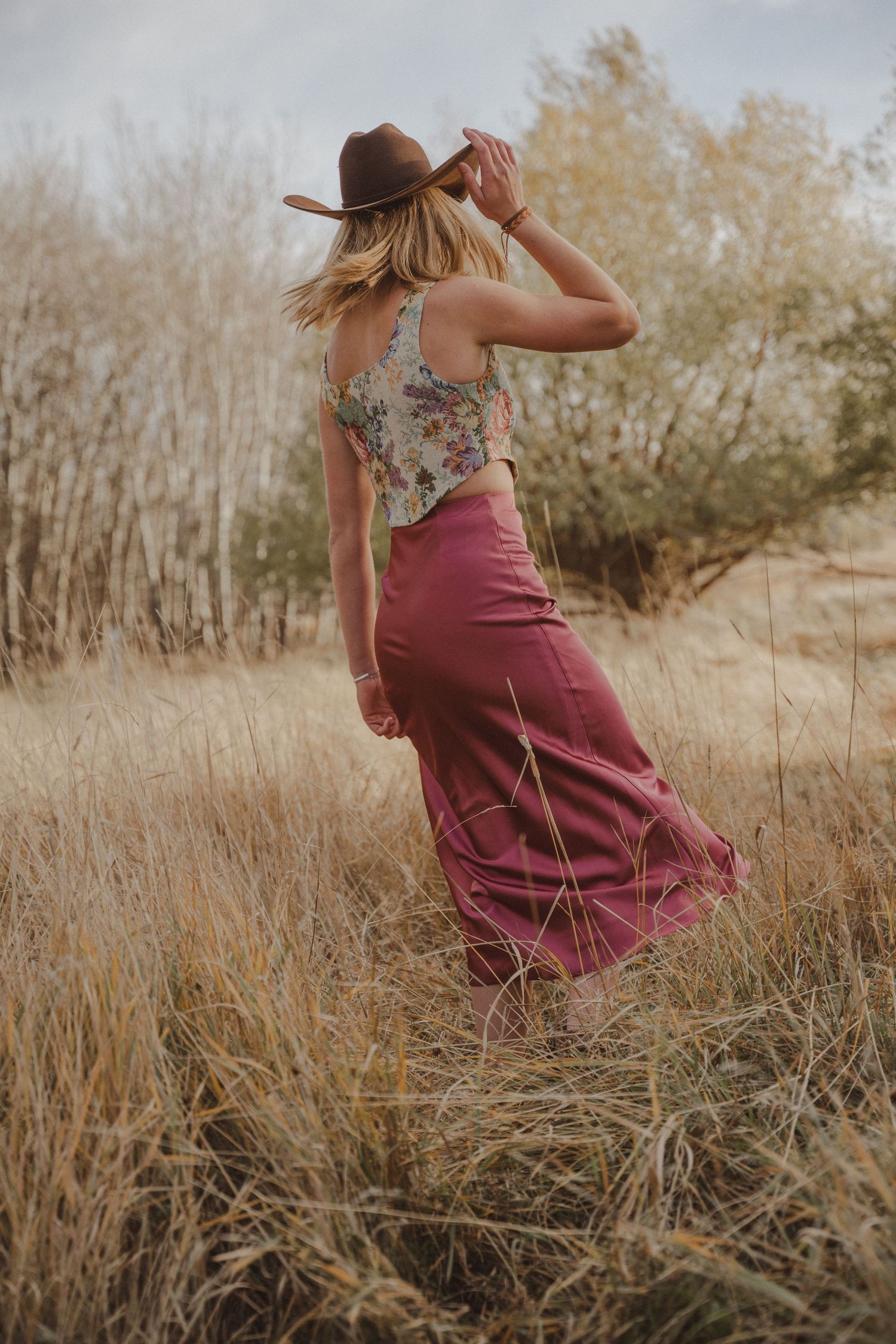 Cowgirl in pink skirt walking through tall grass during golden hour.