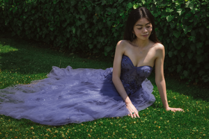 Figure in purple gown lying gracefully on grass with garden backdrop.