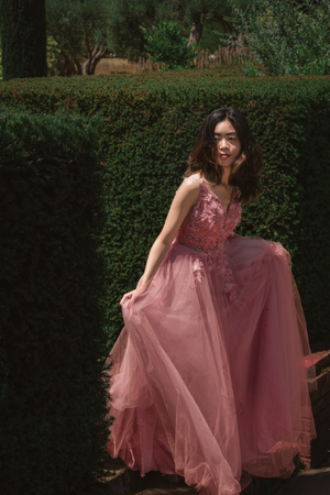 Person in rose-colored tulle gown twirling in garden with hedge backdrop.