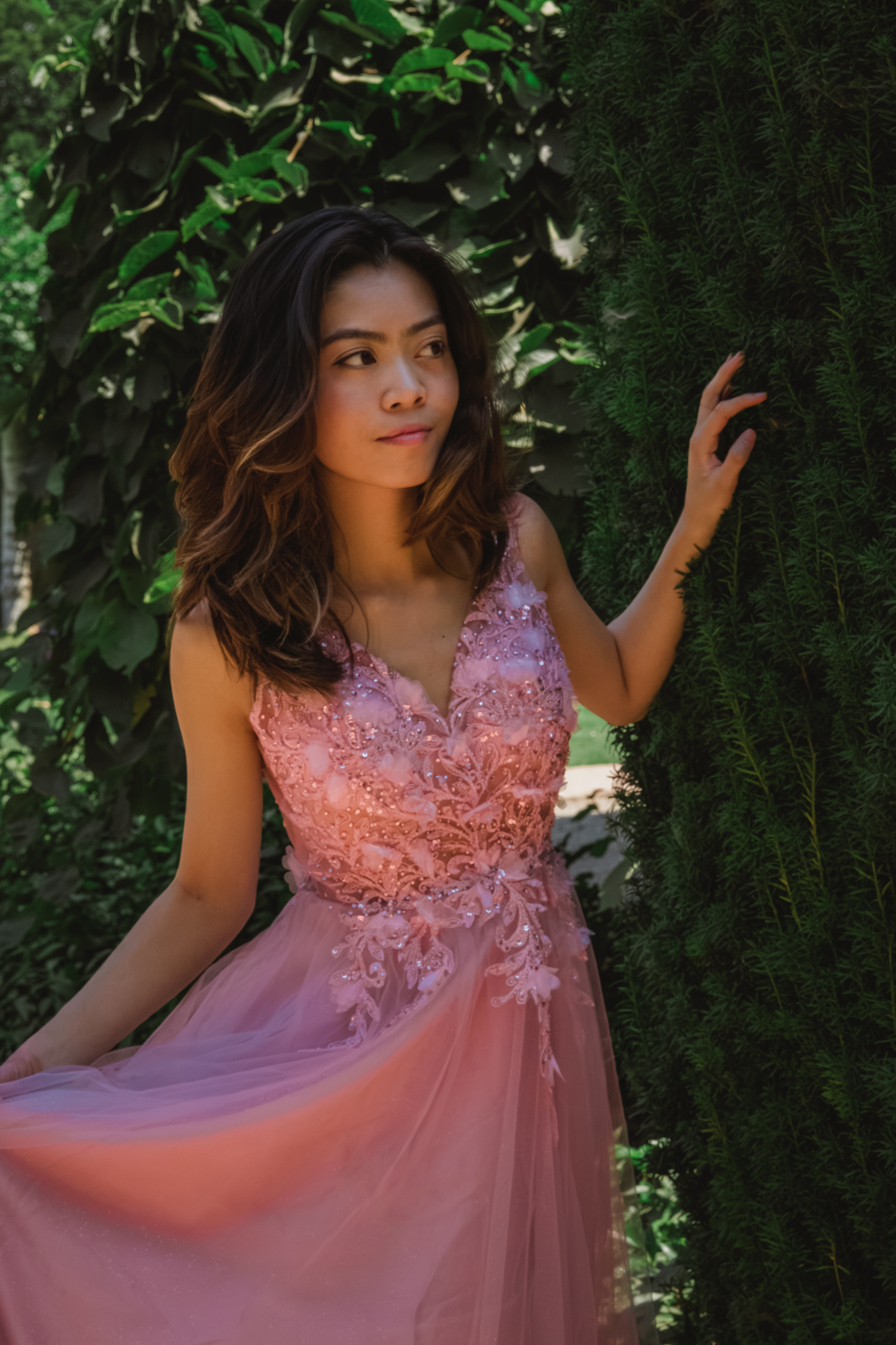 Person in flowing pink lace dress poses against leafy green garden backdrop.