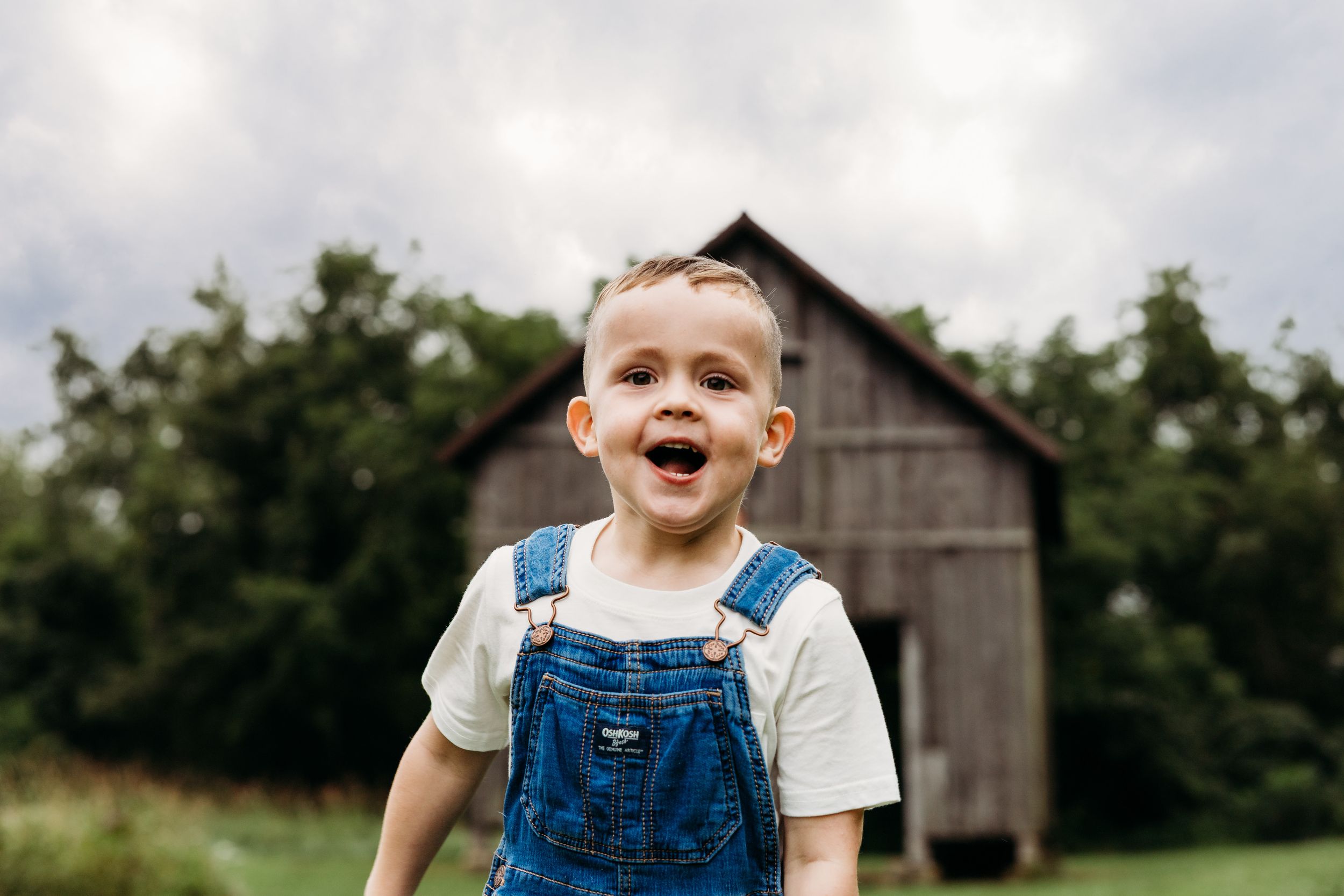 A laughing child in overalls plays outdoors near a rustic barn on a sunny summer day.