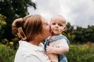 A parent tenderly embraces and kisses a young baby outdoors in a lush natural setting with sunlight streaming through.