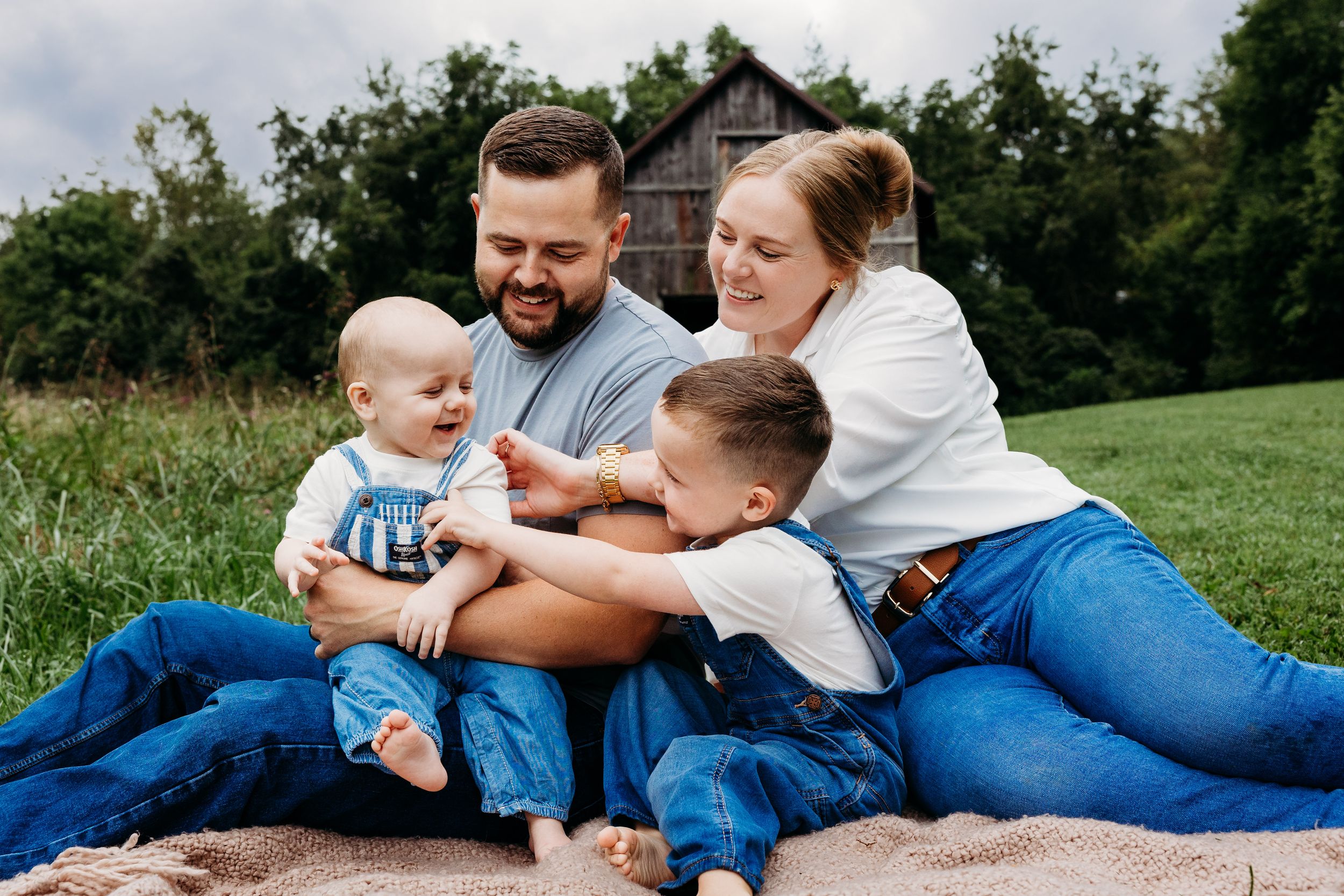 A family wearing jeans and white tops plays together while lying on green grass in a park on a sunny day.