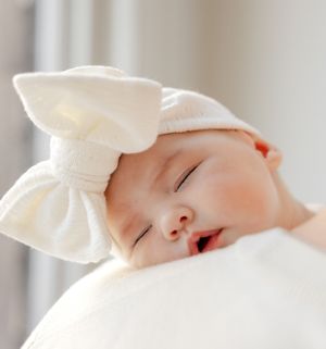 A sleeping newborn baby wrapped in a soft white blanket and hat rests peacefully on a light-colored surface.