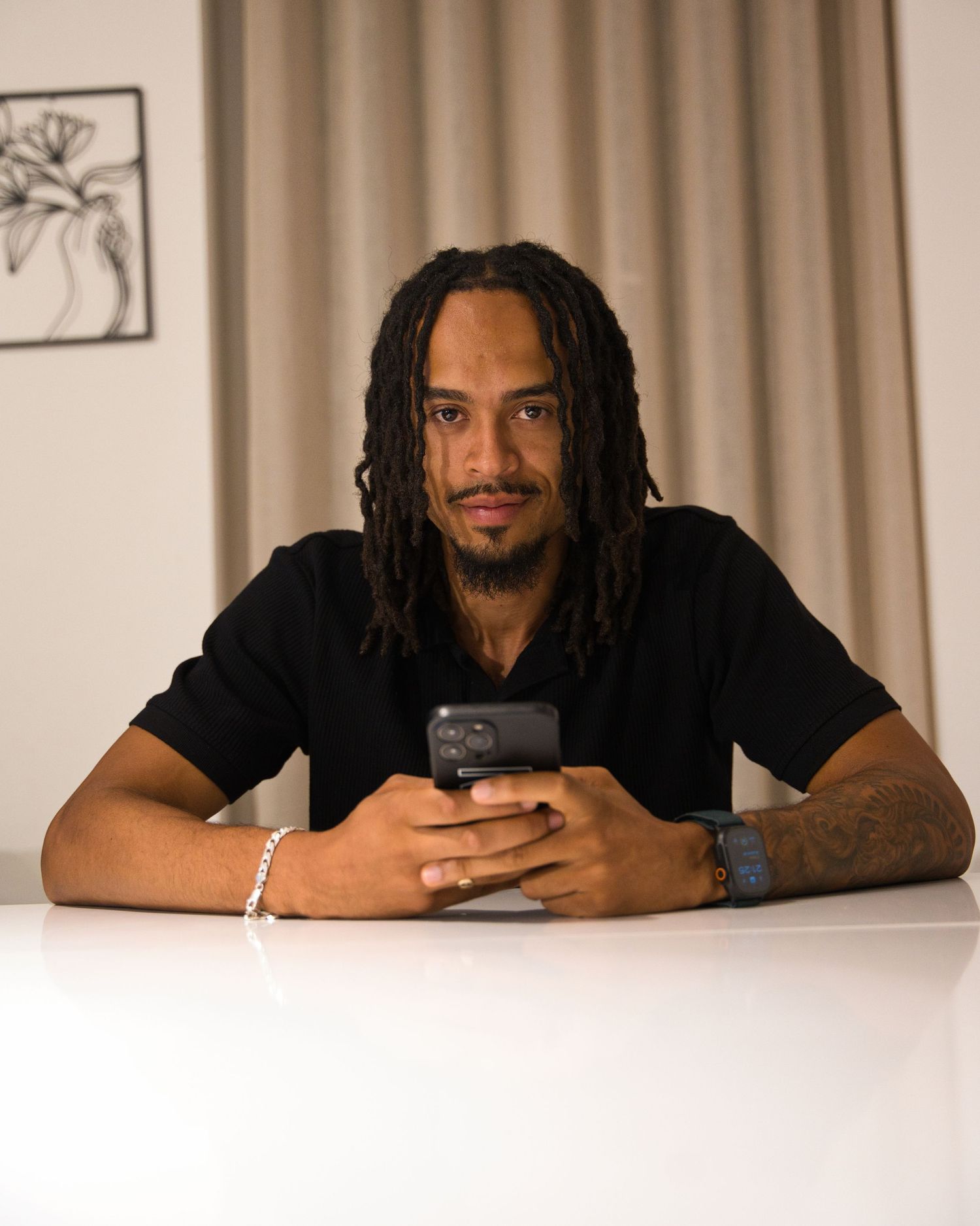 Young man with dreadlocks in casual attire sits looking at phone in warm atmospheric lighting.