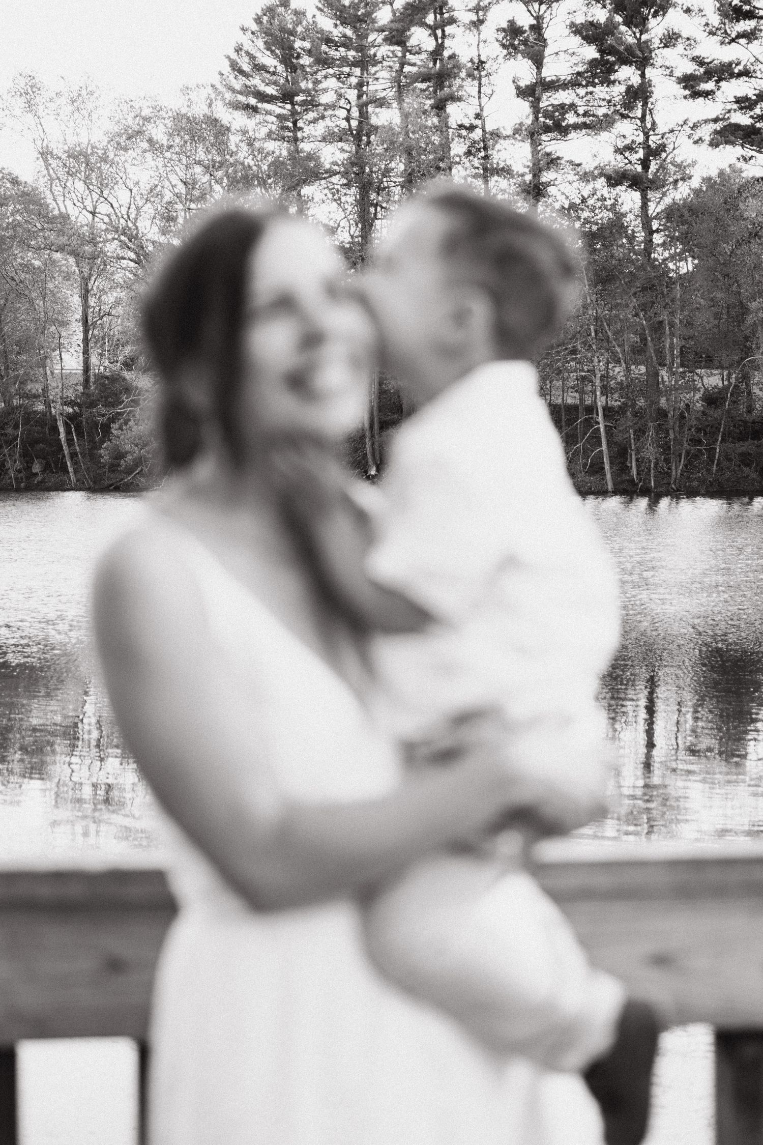 A mother embraces her son while standing on a wooden deck overlooking a serene lake surrounded by pine trees.