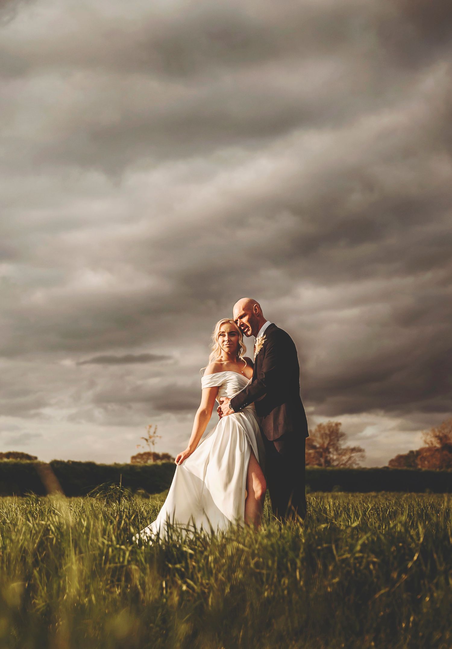 A couple embraces in a grassy field against dramatic stormy clouds at sunset, creating a romantic wedding photo.