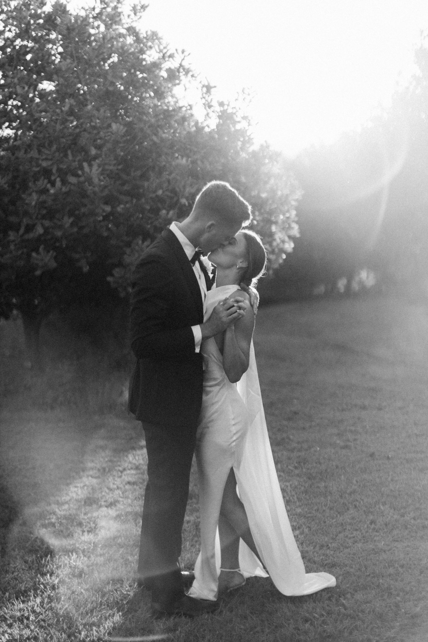A tender romantic outdoor wedding moment captured in black and white as a couple embraces in golden sunlight.