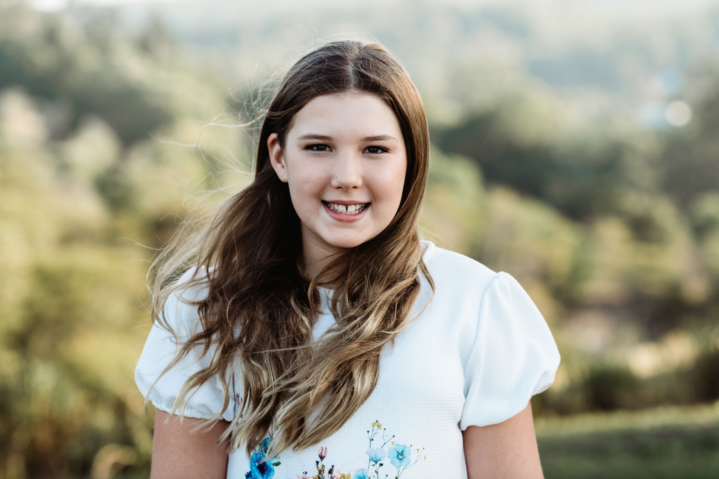 A person wearing a white t-shirt smiles outdoors in natural lighting with mountains in the background.