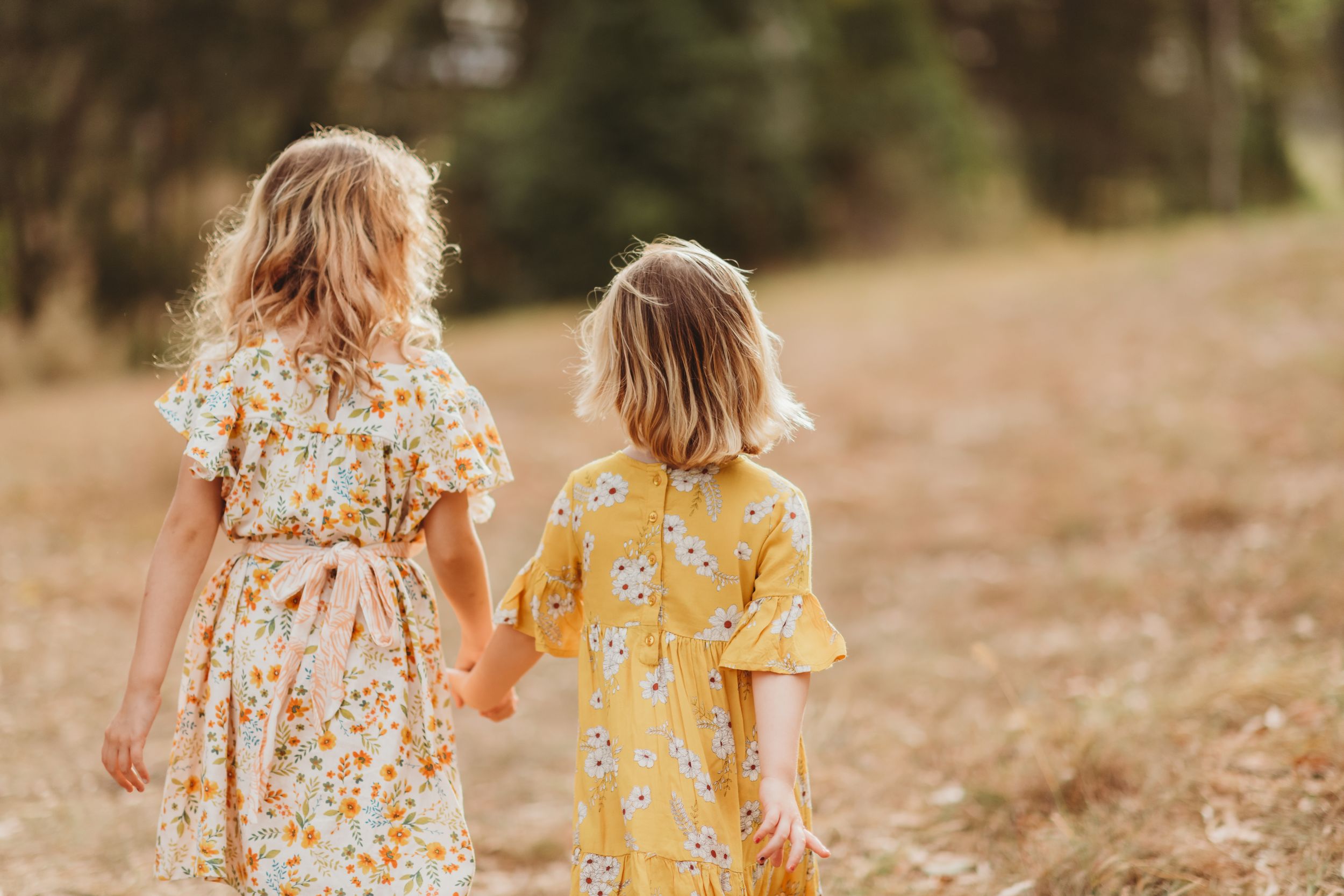 Two children in floral dresses walk hand in hand through a sun-drenched field.