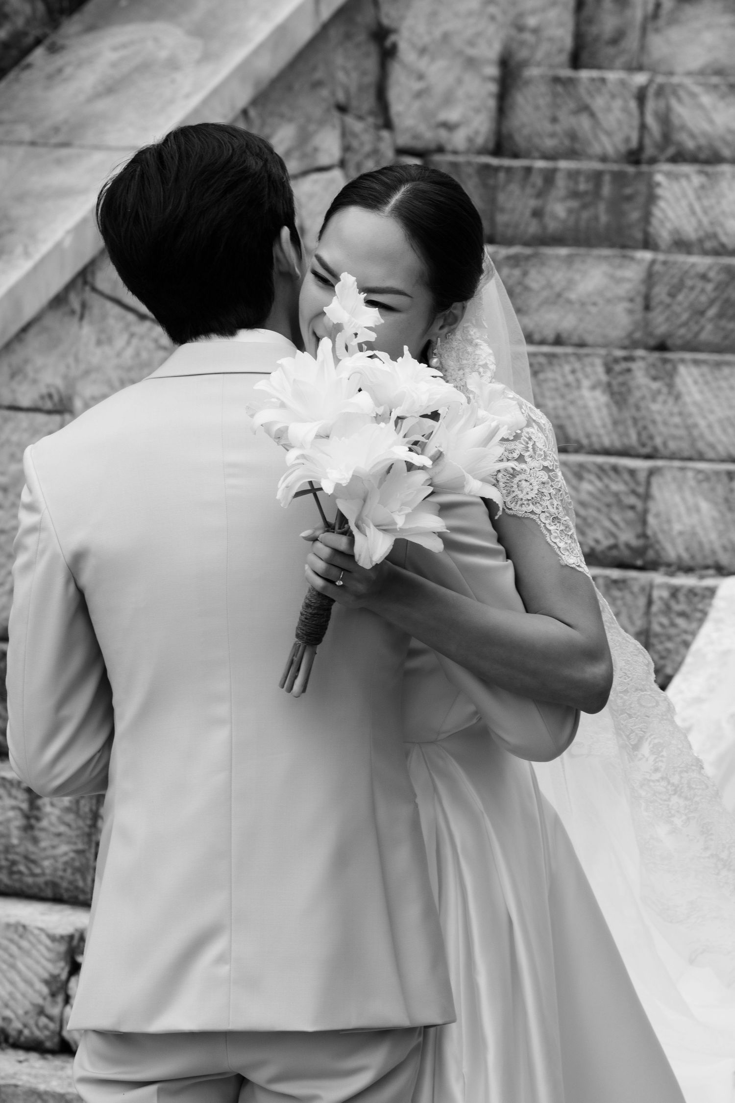Black and white photo of bride and groom embracing with white floral bouquet on stone staircase.
