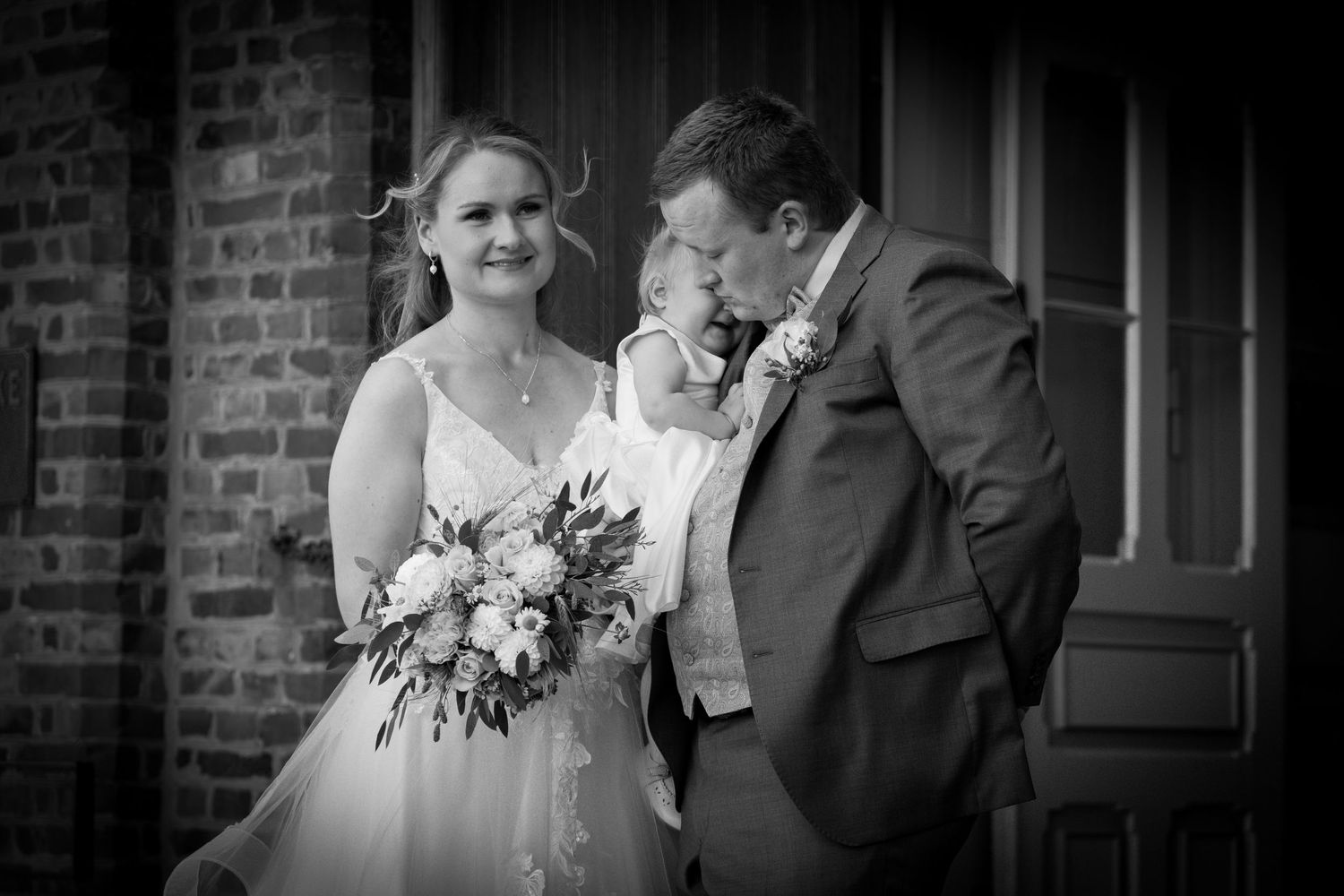 Bride and groom smile with adorable child in black and white wedding portrait outdoors.