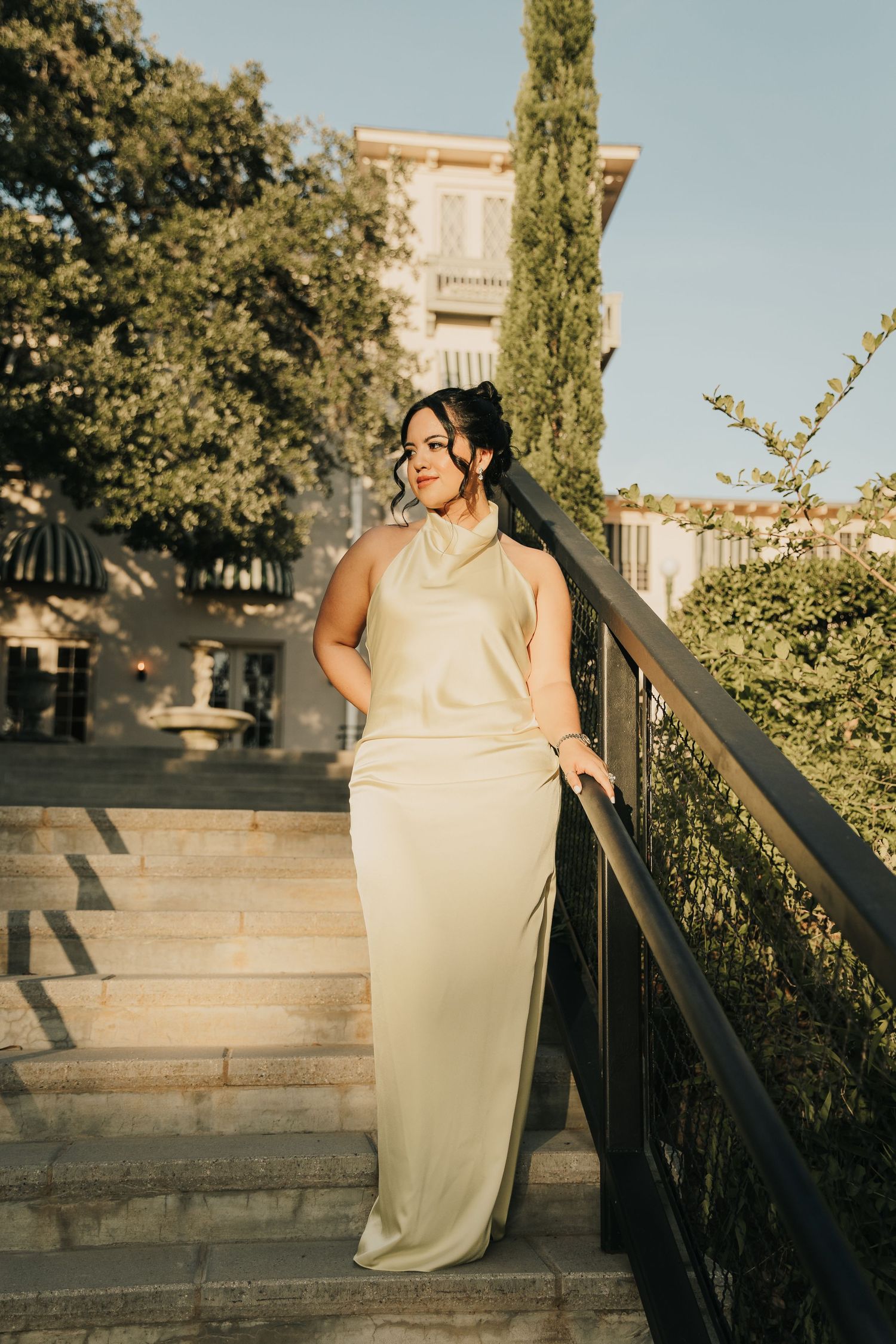 Elegant cream formal dress glows in golden sunset light on stone steps with cypress trees and mansion in background.