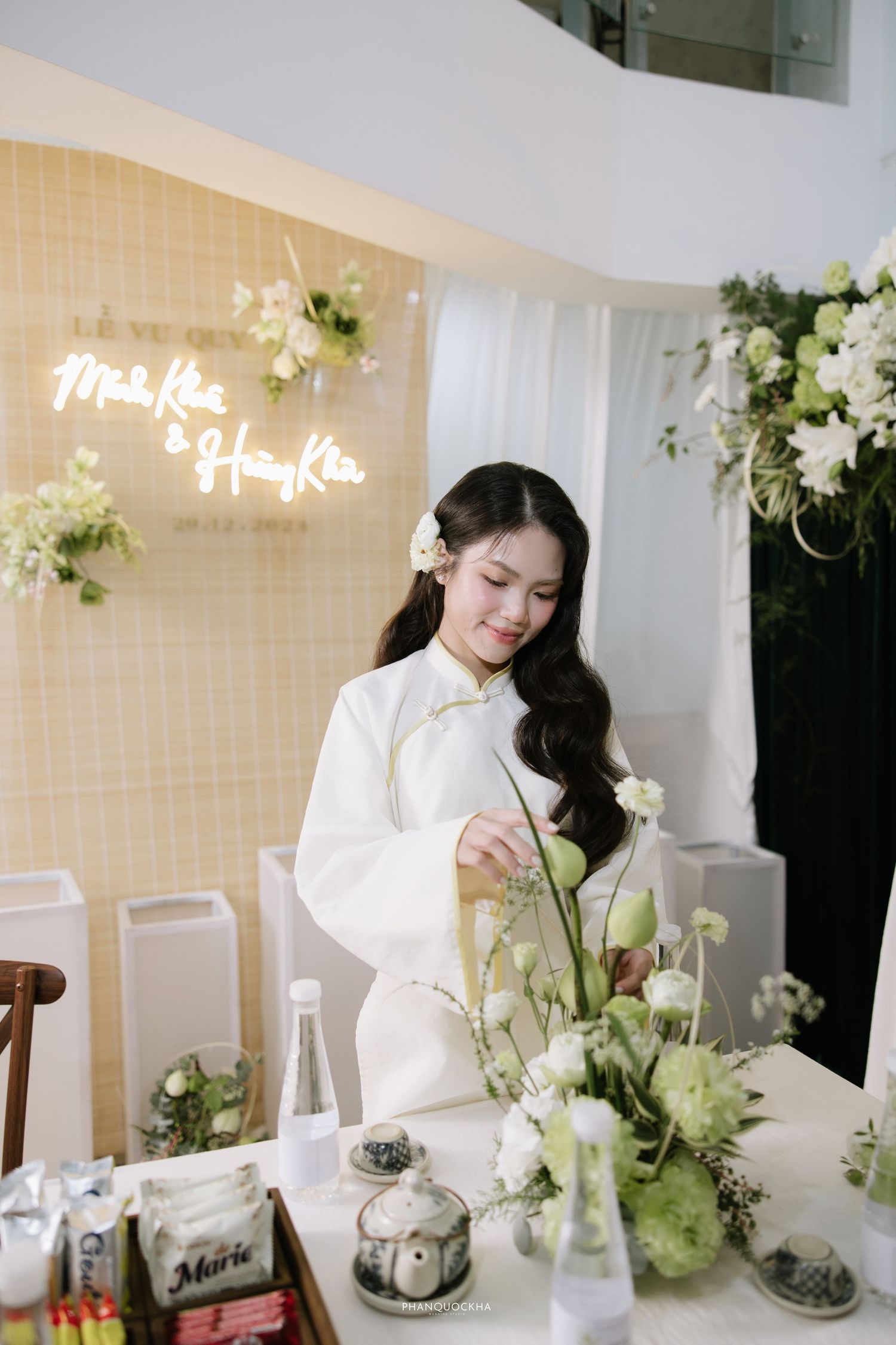 Young woman in white ao dai arranges green and white flowers at decorated Vietnamese wedding ceremony.