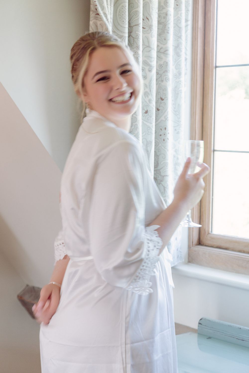 Someone wearing a white robe smiles while standing near a window with curtains in a bright, sunlit room.