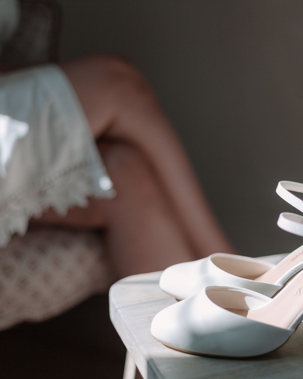 White satin wedding heels with bow detail rest elegantly on a dark surface near lace fabric.