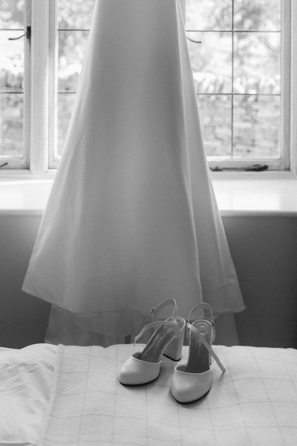 Black and white photo of elegant white wedding dress and bridal shoes near a sunlit window.