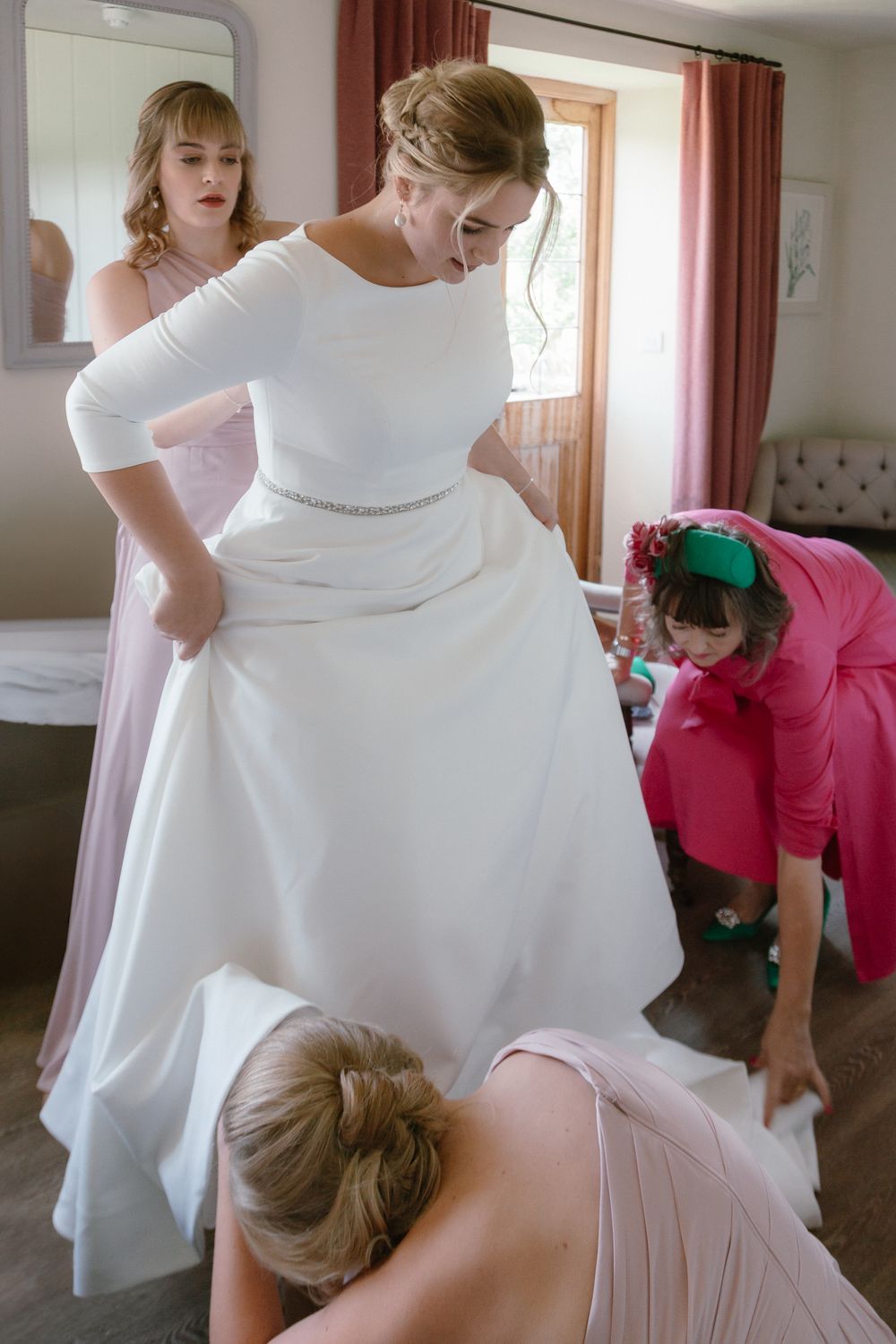 A bride gets help putting on her elegant white wedding gown as others assist with final adjustments in a sunlit room.